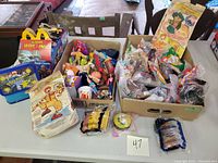 Photo showing two boxes filled with various loose McDonald's Happy Meal toys and collectibles, some in original packaging, on a table.