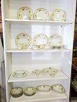 Wide view of Noritake Acacia china set displayed on white shelving including plates, tea cups, serving dishes.