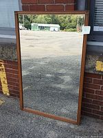 Full view of the large rectangular vintage wall hanging mirror standing against a brick wall, showing reflective glass and wooden frame.