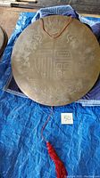 Front view of brass gong showing full etched designs and red hanging string with tassel