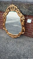 Photo of vintage gold gilt wall mirror leaning against a brick wall, showing the ornate carved floral and leaf details on top and sides of the frame.