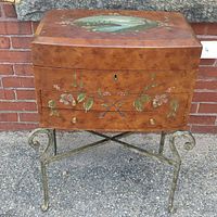 Full view of vintage wooden vanity chest on cast iron stand, showing curved lid with landscape painting and floral details on front drawer.