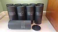 Set of 10 black Riedel branded metal cylindrical canisters for wine glasses with removable lids, lined up on counter.