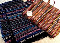 Photo of two colorful woven textiles folded on a bed or flat surface showing intricate geometric, zigzag and stripe patterns in dark blue, purple, black and multi-colored warm tones.
