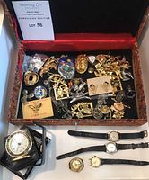 Overhead view of open decorative box showing assorted brooches, pins, pendants and front display of wristwatches and travel clock