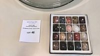 Photo of the 20 carved mineral and gemstone eggs arranged in a white box with individual compartments. An auction lot card is visible nearby.
