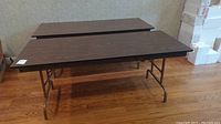 Two 6-foot folding tables side by side, showing dark wood-grain melamine tops and metal legs on wooden floor