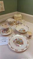 Seven vintage Royal Doulton Bunnykins dishes including mugs, plates, bowl, and small cup displayed on counter with auction lot card.