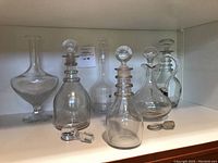 Five glass decanters arranged in a row with clear stopper placed in front, showing variety of shapes and styles including one with ribbed neck and two with smooth bodies