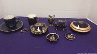 Photo shows full collection of small dark blue and gold miniature porcelain items on purple cloth background. Includes cups, plates, bowls, covered dish, trinket box, and pitcher.