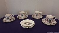 Four coffee cups and saucers arranged with three shell-shaped plates on purple fabric background showing playing card designs.