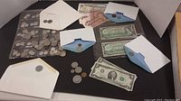 Overview of coins and paper money on black background with several white and blue envelopes containing coins, two Canadian $1 bills, US $1 and $2 bills in view.