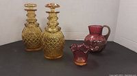 Set of all four glass items: two amber cut glass bottles with lids, cranberry glass pitcher with handle, and small cranberry ruffled edge vase.