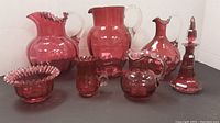 Seven cranberry glass pieces including three pitchers, creamer, bowl, vase, and candleholder