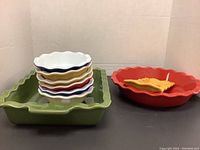 Front view of stacked small ceramic bowls in assorted colors nested inside a large green rectangular casserole dish and next to a red round casserole dish with a small yellow tray on top.