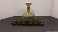 Photo showing the green glass decanter with cork lid centered, with six matching green shooter glasses arranged in front.