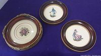 Three framed petit point artworks laid flat on purple fabric background: central floral design in ornate round frame, two smaller round frames with embroidered figures on either side.