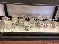 A shelf displaying 14 assorted Portmeirion Botanic Garden mugs featuring detailed botanical floral designs and two matching saucers with green leaf borders.