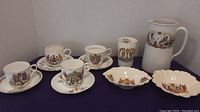 Photo showing a large mug, four tea cups with saucers, and three small decorative dishes, all adorned with British royal portraits and heraldic emblems.