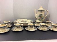 Full coffee set displayed on a black surface with 8 cups and saucers in front, 6 plates stacked behind, and coffee pot on the right side