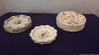 Three stacks of side plates on black cloth background; one smaller stack has 3 Royal Albert plates with gold scalloped edges, right stack has 8 Spode plates with floral prints, left stack has 6 Grimwade octagonal plates with bird and floral design.