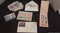 Photo showing multiple sheets of collector stamps in plastic sleeves, an envelope with stamps visible, and a small album containing various stamps laid out on a black surface.