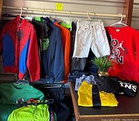 Full view of boys clothing hanging and folded on table showing variety of hoodies, t-shirts, shorts and colors.