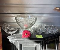 Wide view of the full lot showing the large punch bowl with hobnail pattern, smaller hobnail bowl, and set of small cups arranged on a shelf with a red decorative flower in front. The punch bowl and glasses are clear glass with vintage wear.