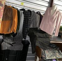 Multiple ladies jackets and a floral carry bag arranged on hangers and furniture, showing variety of styles and colors