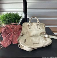 Pink snakeskin-textured crossbody bag next to cream leather bag on a black table with plant background.