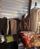 First image showing hanging suede and corduroy pants, brown satin blouse, tan dress on mannequin, multi-color scarf and other pieces laid on wood surface.