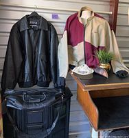 Lot display showing 3XLT black leather jacket on left, color-block beige and burgundy jacket with Aeropostale cap on small table right, and black Canyon Outback leather bag below jackets.