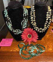 Two natural stone multi-strand necklaces on black necklace display stands, one with pale green stones, the other with creamy white stones. Below is a pink flower and two multi-strand beaded bracelets in turquoise and dark green.