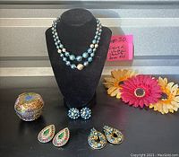 Necklace and various earrings arranged on black jewelry bust and surface, with cloisonné apple-shaped trinket box and some fabric flowers in background