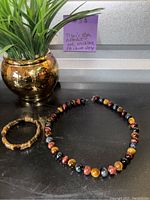 Set displayed on black surface beside a gold-tone round metal planter with green artificial plant. Includes Tiger's Eye beaded necklace and matching bracelet.