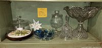 Shelf with vintage glassware set including pedestal bowl, glass bell shell dish with gems, painted tile dome server with wooden base, and smaller glass containers.