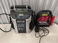 Shows both battery packs side by side, featuring Eliminator pack with handle, air compressor, digital display, and USB ports beside the black and red MotoMaster pack with attached cables.