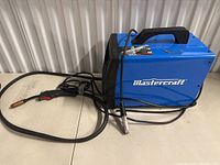 Blue Mastercraft welding machine with attached welding gun and cable laid on table.