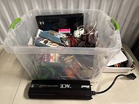 Clear storage bin filled with various electronic cables, adapters, and components with car power inverter in front.