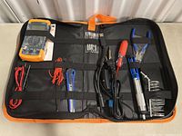 View of entire tool kit in black and orange case including multimeter, screwdrivers, soldering iron, test leads, wire cutters, tweezers, wrenches.