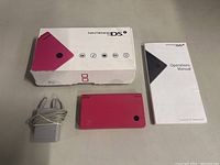 Box with pink Nintendo DSi console, gray charger, and white operations manual on a flat surface.