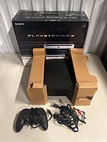 Sony PlayStation 3 60GB console with one black wired controller and AV cables, placed in front of the original box.