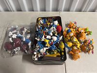 Top view showing Raisins characters in a plastic bag, Smurfs in a tin box, and Garfield figurines adjacent on a white table.
