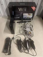 Full view of Nintendo Wii console standing upright with original box behind and accessories laid out in front including remote, Nunchuks, sensor bar, and AV cable.