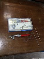 Two photos showing a Paasche airbrush kit. First photo shows the kit case, airbrush and tools on wood surface. Second photo zooms in on kit contents and loose airbrush below.