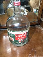 Full view of vintage Coca Cola one gallon glass syrup jug with embossed text, paper label, and screw cap.