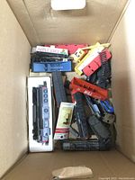 Overhead view of cardboard box containing various HO scale train cars and engines in assorted colors including red, yellow, blue, and gray. Some cars are partially assembled and in various states of condition.