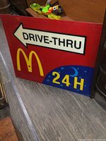 Red rectangular McDonald's plexiglass sign featuring a white arrow pointing left with 'DRIVE-THRU' text in black, golden arches logo, and 24H blue moon symbol.