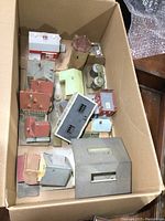 Box containing multiple model railroad structures arranged compactly within a cardboard box. Visible are various roofs and small building parts in diverse colors including red, brown, grey, green and white.