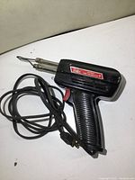 View of the soldering tool showing the dual soldering tips, black handle with ribbed grip, power cord, and red trigger switch, against a white background.